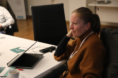 Emilia sitting at a table with a laptop, listening in a meeting